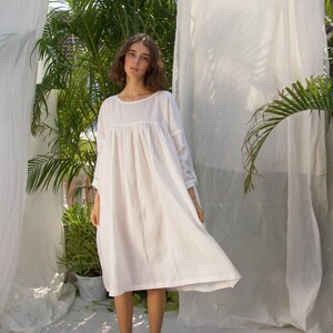 Puede incluir: Vestido de lino blanco con cuello redondo y mangas largas. El vestido tiene una silueta suelta y fluida y llega hasta la pantorrilla. La modelo est&aacute; de pie frente a tela blanca y vegetaci&oacute;n.
