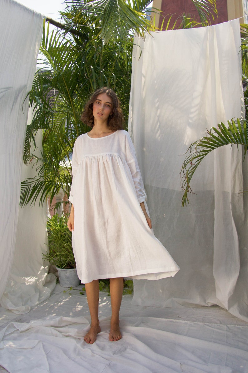 Puede incluir: Vestido de lino blanco con cuello redondo y mangas largas. El vestido tiene una silueta suelta y fluida y llega hasta la pantorrilla. La modelo est&aacute; de pie frente a tela blanca y vegetaci&oacute;n.
