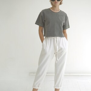 May include: A woman wearing a gray cropped t-shirt with white stitching and white pants with a drawstring waist. She is wearing black sunglasses and a pearl necklace.