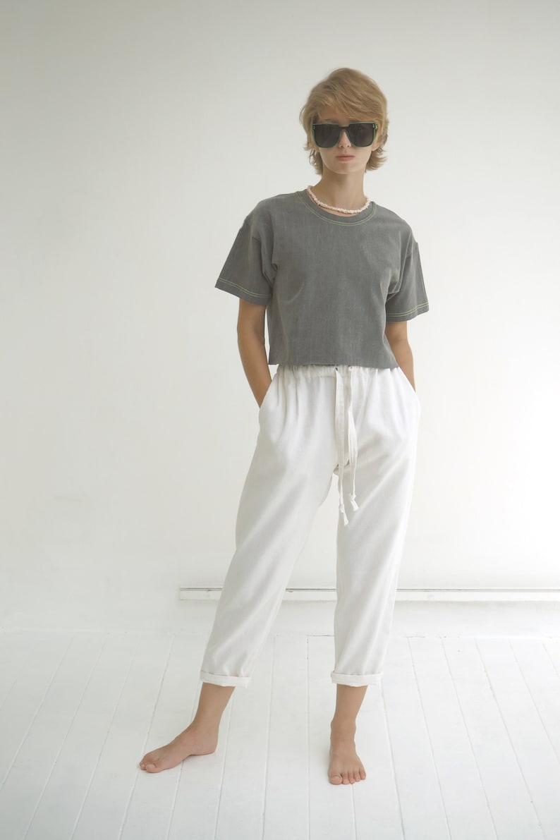 May include: A woman wearing a gray cropped t-shirt with white stitching and white pants with a drawstring waist. She is wearing black sunglasses and a pearl necklace.