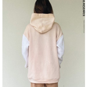 May include: A beige and white colour-block hoodie with a drawstring hood. The sweatshirt has white sleeves and a beige body and hood. The text "TROPICAL RESEARCH" is printed vertically on the right side. The text "THE SWEAT-SHIRT" is printed on the bottom left.