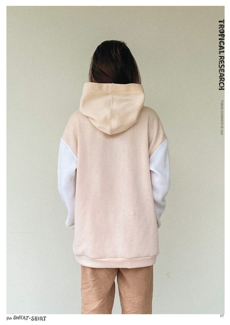 May include: A beige and white colour-block hoodie with a drawstring hood. The sweatshirt has white sleeves and a beige body and hood. The text "TROPICAL RESEARCH" is printed vertically on the right side. The text "THE SWEAT-SHIRT" is printed on the bottom left.