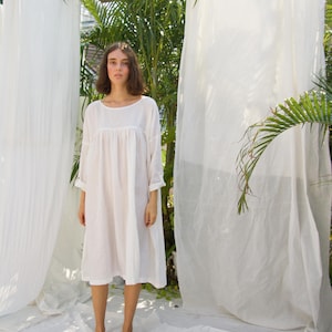 Puede incluir: Vestido blanco holgado, con cuello redondo y mangas tres cuartos. El vestido llega a la rodilla y est&aacute; hecho de una tela ligera. La modelo est&aacute; de pie frente a tela blanca y vegetaci&oacute;n.