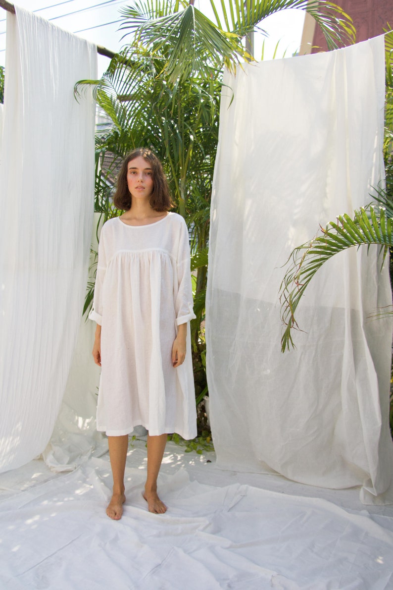Puede incluir: Vestido blanco holgado, con cuello redondo y mangas tres cuartos. El vestido llega a la rodilla y est&aacute; hecho de una tela ligera. La modelo est&aacute; de pie frente a tela blanca y vegetaci&oacute;n.