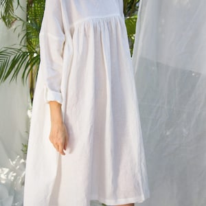 Puede incluir: Vestido blanco de lino con una silueta suelta y fluida. El vestido presenta un cuello redondo, mangas largas con pu&ntilde;os enrollados y un corpi&ntilde;o fruncido. El dobladillo cae por debajo de las rodillas. Hecho de tela ligera.