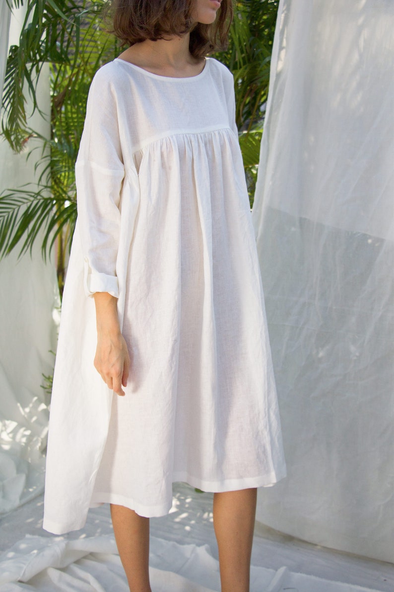 Puede incluir: Vestido blanco de lino con una silueta suelta y fluida. El vestido presenta un cuello redondo, mangas largas con pu&ntilde;os enrollados y un corpi&ntilde;o fruncido. El dobladillo cae por debajo de las rodillas. Hecho de tela ligera.