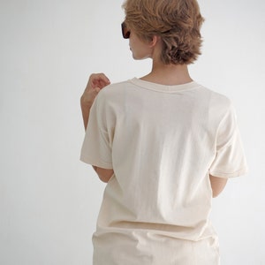 May include: A woman wearing a light beige t-shirt with short sleeves and white pants. She is wearing sunglasses and has short, light brown hair.