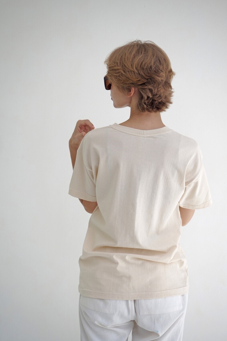 May include: A woman wearing a light beige t-shirt with short sleeves and white pants. She is wearing sunglasses and has short, light brown hair.