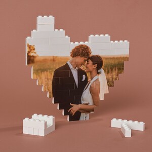 May include: A white LEGO heart-shaped frame with a photo of a couple in wedding attire. The couple is looking at each other and smiling. The photo is set against a backdrop of a field with tall grass and a sunset.