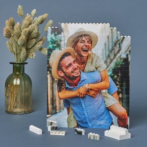 May include: A couple smiles for a photo while standing in a brick alleyway. The man is wearing a blue shirt and the woman is wearing a white dress. The photo is printed on a white background and is made of small plastic bricks.