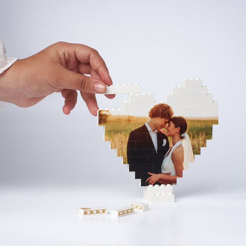 May include: A white heart-shaped frame made of Lego bricks with a photo of a bride and groom inside. The photo shows the couple looking at each other and smiling. There are several white Lego bricks scattered around the frame.