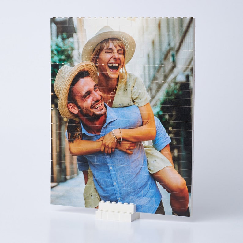 May include: A couple is laughing and smiling while the man carries the woman on his back. The woman is wearing a white dress and a straw hat. The man is wearing a blue shirt and a straw hat. They are both looking at the camera.