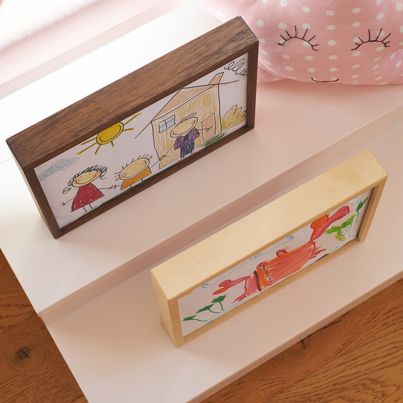 Puede incluir: Dos dibujos de ni&ntilde;os enmarcados en un estante blanco. El dibujo superior est&aacute; en un marco marr&oacute;n y representa a una familia de tres con una casa y un sol. El dibujo inferior est&aacute; en un marco de madera clara y muestra un cangrejo rojo.