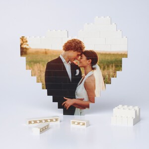 May include: A white Lego heart-shaped frame with a photo of a bride and groom inside. The couple is looking at each other and smiling. There are also some loose Lego bricks on the table.