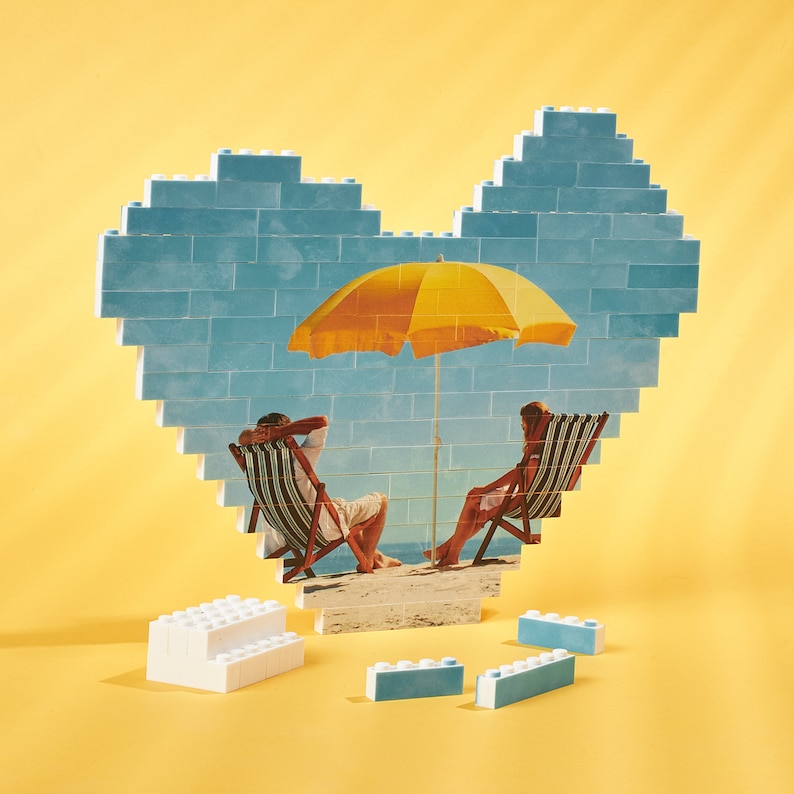 May include: A blue heart-shaped brick wall with a yellow umbrella, two chairs, and two people sitting in the chairs. The scene is a beach with sand and blue sky.