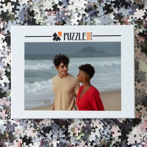 May include: A 500 piece jigsaw puzzle featuring a couple standing on a beach. The man is wearing a beige shirt and the woman is wearing a red dress. The ocean is in the background.