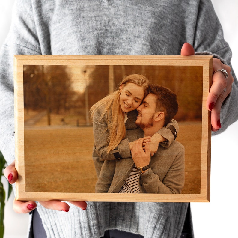 Puede incluir: Un marco de madera clara con una foto de una pareja abraz&aacute;ndose y sonriendo. La foto tiene tonos c&aacute;lidos. La pareja lleva abrigos grises. El marco es de madera clara.