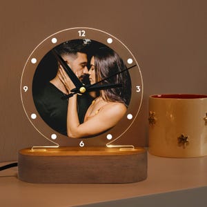 May include: A wooden base with a clear acrylic clock face featuring a couple's photo. The clock has black hands and white numbers. The base has a warm light that illuminates the clock face.
