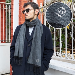 Op de afbeelding: Een grijze sjaal met visgraatpatroon en zwarte franjes. De sjaal is om de nek van een persoon gewikkeld en heeft de initialen "N.D." in een cirkelvormig ontwerp. De persoon draagt een zonnebril en een donkere jas.