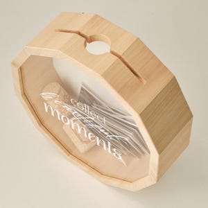 May include: Wooden box with a glass front and a slot on the top. The box is filled with cork stoppers and the text "Collect beautiful moments" is printed on the glass.