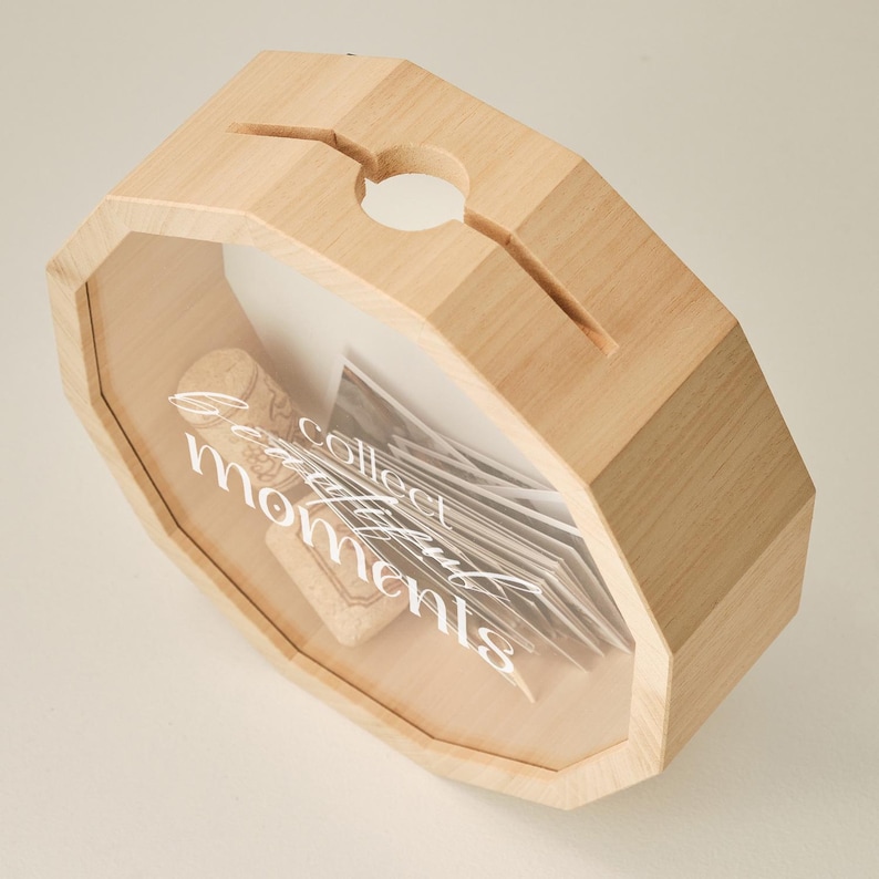 May include: Wooden box with a glass front and a slot on the top. The box is filled with cork stoppers and the text "Collect beautiful moments" is printed on the glass.