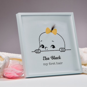 May include: A white frame containing a black and white illustration of a baby peeking over a surface. The baby has a yellow bow in its hair. The text "Elsa Black my first hair" is printed below the illustration.