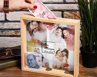 Op de afbeelding: Een houten fotolijst met een collage van foto's van een stel. De lijst heeft een gleuf om geld in te stoppen en is gepersonaliseerd met de namen "Mia Morgan" en de datum "12.12.2018".