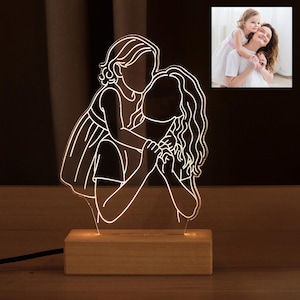 May include: A white outline of a mother and child hugging, illuminated by a light source from below. The light is on a wooden base.