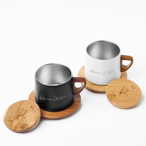 May include: Two stainless steel mugs, one black and one white, with wooden handles and the name "Raina Wilson" printed on the side. They sit on wooden coasters, each with a lid featuring an "R" and starburst design.