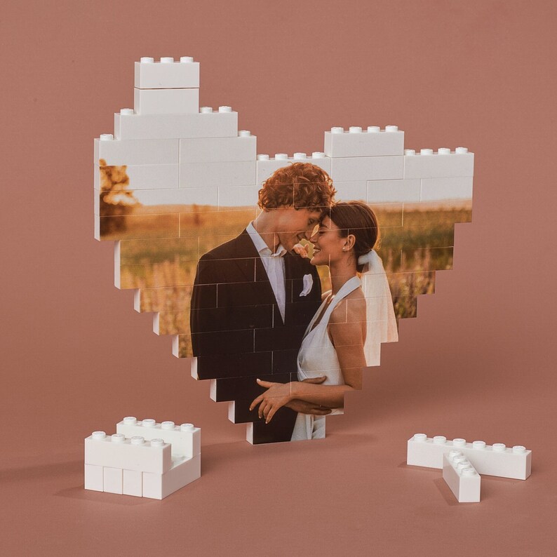 May include: A white LEGO heart-shaped frame with a photo of a couple in wedding attire. The couple is looking at each other and smiling. The photo is set against a backdrop of a field with tall grass and a sunset.