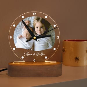 Może przedstawiać: Drewniana podstawa z przezroczystym akrylowym cyferblatem z fotografią dwójki dzieci. Cyferblat ma czarne wskazówki i białe cyfry. Podstawa ma światło, które oświetla cyferblat. Na cyferblacie widnieje napis "Sera & Peter".