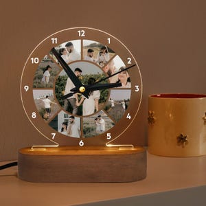 Puede incluir: Una base de madera con una esfera de reloj de acrílico transparente que presenta 12 ranuras para fotos. La esfera del reloj tiene una manecilla negra y detalles dorados. El reloj está iluminado con luces LED de color blanco cálido.