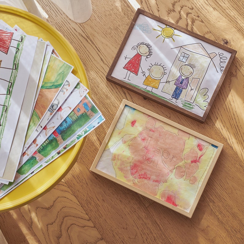 Puede incluir: Tres dibujos enmarcados sobre una superficie de madera. El primer dibujo es una familia de tres figuras de palo con una casa y un sol. El segundo dibujo es una pintura de acuarela con las palabras "Te amo" en rosa y rojo. El tercer dibujo es una pila de papeles con varios dibujos en ellos.