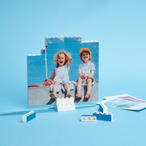 May include: A photo puzzle featuring two children sitting on a ledge. The girl is wearing a white shirt and has long blonde hair. The boy is wearing a white shirt and a yellow baseball cap. The puzzle is made of white plastic bricks.