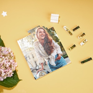 May include: A photo puzzle featuring a woman with long brown hair smiling and wearing a white shirt and blue jeans. The puzzle is made up of small white squares.
