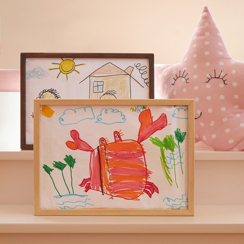 Puede incluir: Dos dibujos de ni&ntilde;os enmarcados. El dibujo de arriba muestra una casa, un sol y dos personas. El dibujo de abajo muestra un cangrejo rojo con rayas naranjas, plantas verdes y olas azules. La palabra "mira" est&aacute; escrita en la esquina superior derecha del dibujo de abajo.