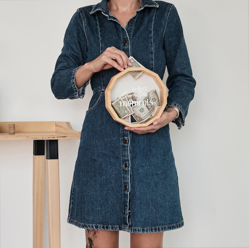 May include: A wooden money box with a clear glass top and the text "collect moments" printed on the front. The box is filled with US dollar bills. A person is holding the box in their hands.