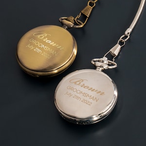 Könnte beinhalten: Zwei goldene Taschenuhren mit einer Kette. Die Zifferblätter der Uhren sind mit dem Text "Brown, Groomsman, July 21th 2022" graviert.