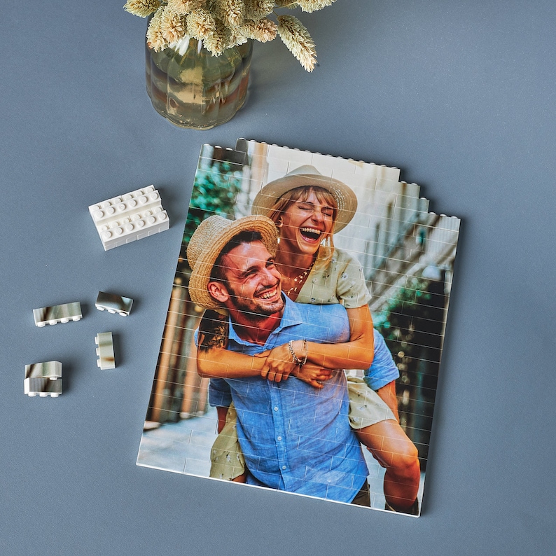 May include: A couple poses for a photo. The woman is wearing a white dress and a straw hat. The man is wearing a blue shirt and a straw hat. They are standing in front of a brick wall.