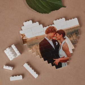 May include: A white LEGO heart-shaped frame with a photo of a couple kissing. The couple is dressed in formal attire and is standing in a field.