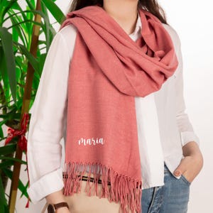 May include: A coral-colored scarf with fringe detail and the name "Maria" in white script. The scarf is draped over a white shirt and blue jeans. The scarf is made of a soft material.