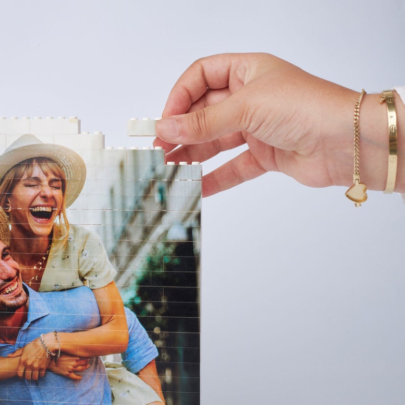 May include: A couple poses for a photo while putting together a white jigsaw puzzle. The puzzle features a photo of the couple. The woman is wearing a white hat and a white shirt. The man is wearing a blue shirt.