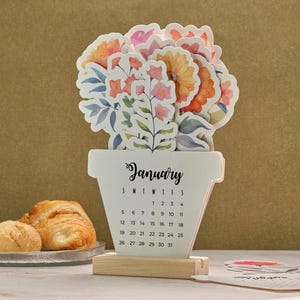 May include: A decorative wooden calendar for January, shaped like a flower pot, with colorful floral cutouts. The calendar displays the days of the month and is supported by a wooden base. The text "January" is written in cursive.