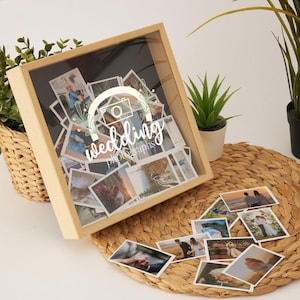 Könnte beinhalten: Eine Holz-Schattenbox mit einer transparenten Vorderseite, gefüllt mit Hochzeitsfotos. Auf der Box steht in weißer Schrift "wedding photographs". Mehrere lose Fotos sind auf einer gewebten Matte um die Box herum verstreut.