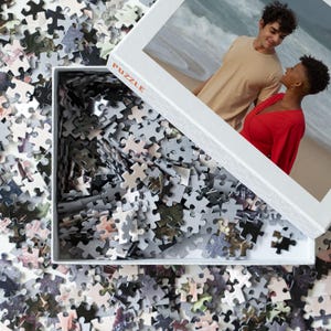 May include: A white box with the word "PUZZLE" printed on it. The box is open and filled with a jumbled pile of puzzle pieces. The image on the puzzle is of a couple standing on a beach.