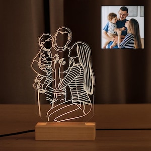 May include: A white outline of a family of three, a man, a woman, and a child, is illuminated on a clear acrylic base. The base is mounted on a wooden stand. The image is lit from behind, creating a warm glow.
