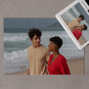 May include: A couple standing on a beach, looking at each other. The man is wearing a beige shirt and the woman is wearing a red dress. The ocean is in the background.