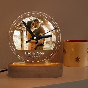 Può includere: Una base in legno con un quadrante dell'orologio in acrilico trasparente che presenta una foto personalizzata di una coppia. Il quadrante dell'orologio ha lancette nere e un bordo dorato. La base ha una luce che illumina il quadrante dell'orologio. Il quadrante dell'orologio include anche il testo "Lisa & Peter" e "04.04.2024".