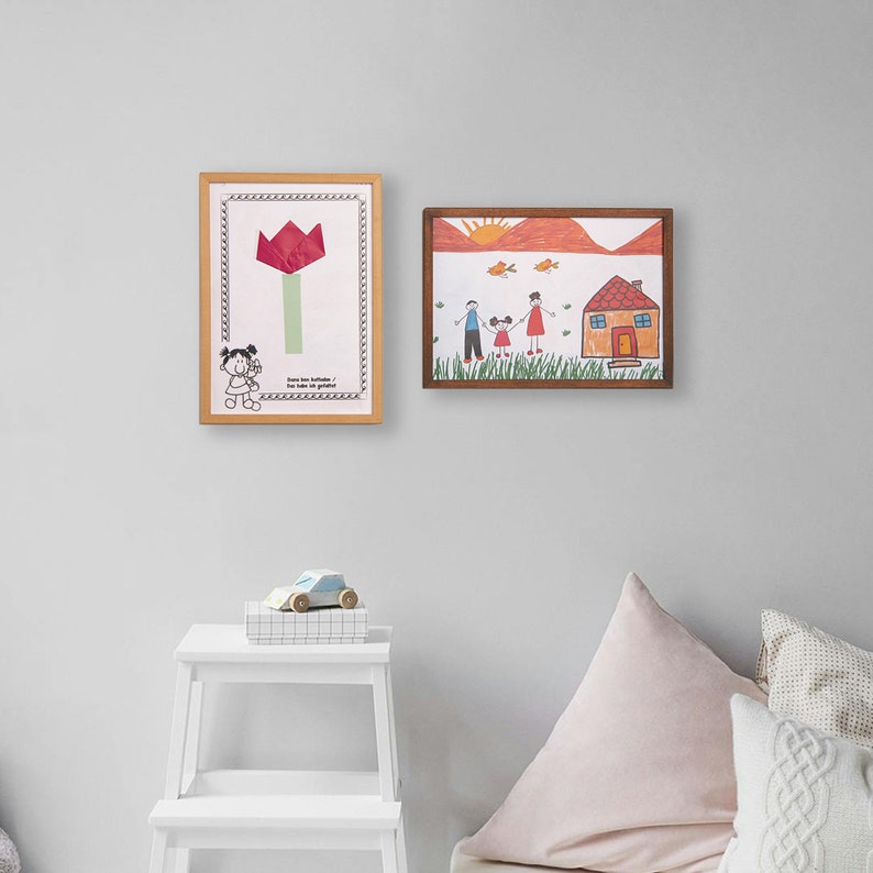 Puede incluir: Dos dibujos enmarcados en una pared blanca. El dibujo de la izquierda es de una flor roja con un tallo verde. El dibujo de la derecha es de una familia de tres personas de pie frente a una casa con un techo rojo. Los dibujos est&aacute;n en un estilo simple, t&iacute;pico del arte infantil.