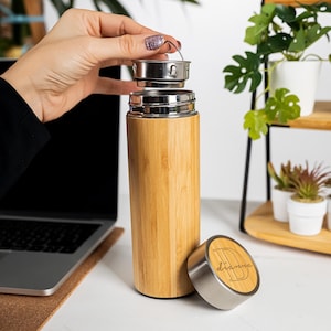 Op de afbeelding: Een bamboe reismok met een roestvrijstalen deksel en een apart deksel met de naam "D'ianna" gegraveerd. De mok is cilindrisch en heeft een natuurlijke houtnerf afwerking. De deksel wordt verwijderd.